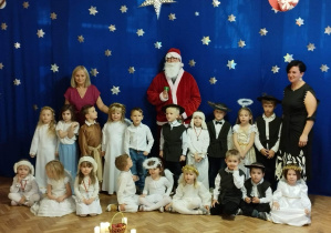 Dzieci pozują do zdjęcia ze św. Mikołajem i swoimi nauczycielkami na tle niebieskiego materiału ze srebrnymi gwiazdkami.