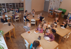 Dzieci siedzą przy stolikach i jedzą własnoręcznie przygotowane pizze.