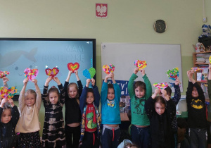 Na zdjęciu dzieci z grupy czerwonej dzieci trzymają serca i stoją w klasie na tle tablicy multimedialnej