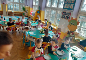 Na zdjęciu dzieci z grupy fioletowej siedzące przy stolikach dzieci ubrane są w fartuchy