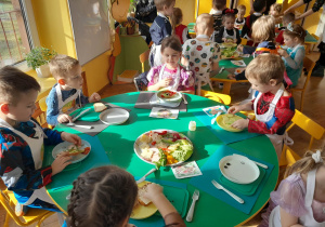 Na zdjęciu dzieci z grupy fioletowej siedzące przy stolikach dzieci ubrane są w fartuchy wykonują kanapki