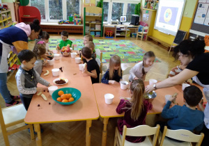 Na zdjęciu dzieci z grupy zielonej siedzą przy połączonych stołach dzieci wykonują sałatki owocowe z pomocą rodziców