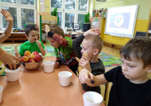 Na zdjęciu czterech chłopców z grupy zielonej siedzący przy stoliku z owocami w ręku chłopcy podnoszą ręce
