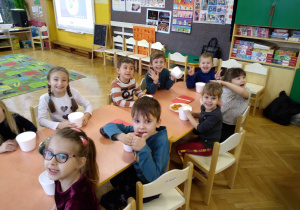 Na zdjęciu dzieci z grupy zielonej siedzą przy połączonych stołach dzieci trzmają w rękach kubeczki na sałatkę dzieci się uśmiechają