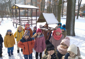 Na zdjęciu dzieci z grupy zielonej ubrane w kurtki stoją w ogrodzie wokół zimowa sceneria za dziećmi karmnik i altana