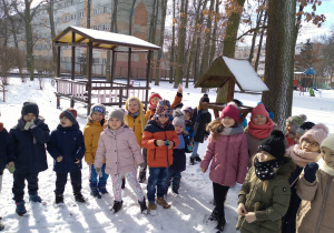 Nazdjęciu dzieci z grupy zielonej stoją w ogrodzie przedszkolnym przy karmniku wokół zimowy krajobraz