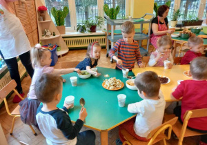 Dzieci siedzą przy stole. Nakładają sobie do kubeczków wybrane, pokrojone przez rodziców i personel przedszkola owoce.