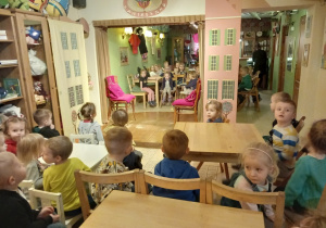 Dzieci siedzą przy stolikach. Czekają na rozpoczęcie przedstawienia. W centralnej części sali znajduje się scena.