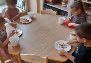 Trzy dziewczynki i jeden chłopiec siedzą przy stoliku. Kroją plastikowymi nożami owoce do sałatki.