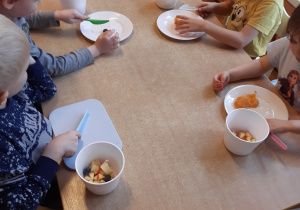 Trzech chłopców i jedna dziewczynka siedzą przy stole. Kroją plastikowymi nożami owoce do swojej sałatki.