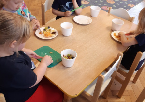 Trzy dziewczynki i jeden chłopiec siedzą przy stoliku. Kroją plastikowymi nożami owoce.
