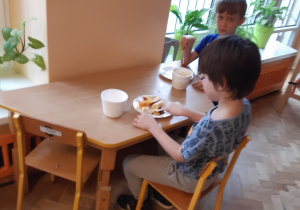 Dwóch chłopców siedząc przy stoliku kroi plastikowymi nożami owoce do sałatki.
