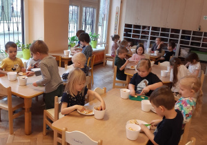 Dzieci z grupy pomarańczowej siedzą przy stolikach. Przygotowują swoją sałatkę owocową- kroją plastikowymi nożami owoce.