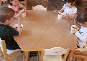 Cztery dziewczynki i dwóch chłopców siedzi przy stolikach. Jedzą własnoręcznie wykonane sałatki owocowe.