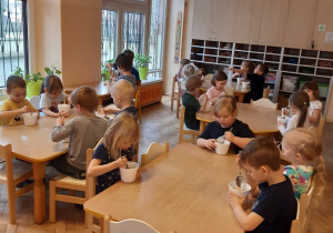 Dzieci z grupy pomarańczowej siedzą przy stolikach. Jedzą własnoręcznie przyrządzone sałatki owocowe.
