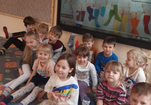 Dzieci siedzą na dywanie w sowy. Na nogach mają założone skarpetki nie do pary. Za dziećmi na tablicy interaktywnej wyświetlona jest główna idea Dnia Zespołu Downa.