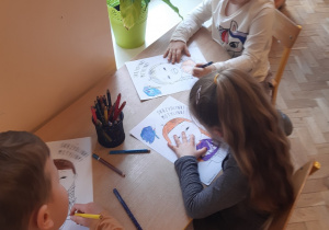 Dwie dziewczynki i jeden chłopiec siedzą przy stoliku- kolorują obrazek przedstawiający dziewczynkę z zespołem Downa.