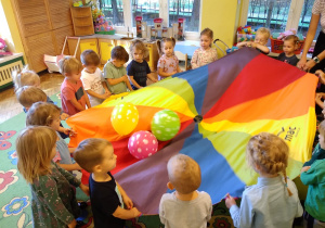 Na zdjęciu dzieci z grupy żółtej stoją na dywanie w kole trzymają kolorową chustę animacyjną, na której znajdują się 3 balony