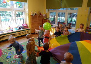 Na zdjęciu dzieci z grupy żółtej stoją na dywanie w kole trzymają kolorową chustę animacyjną, na której znajdują się 3 balony, dzieci poruszają chustą, balony latają w powietrzu