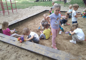 Na zdjęciu dzieci z grupy żółtej podczas pobytu w piaskownicy w ogrodzie dzieci bawią się w piaskownicy jedna z dziewczynek stoi uśmiechnięta