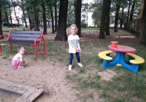 Na zdjęciu dwie dziewczynki bawią się w ogrodzie jedna stoi obok stolika druga siedzi obok ławki