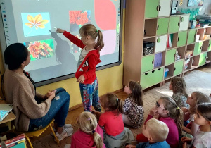 Na zdjęciu dzieci z grupy fioletowej podczas zajęć dzieci siedzą na dywanie i obserwują dziewczynkę, która wskazuje na elementy na tablicy multimedialnej