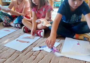 Na zdjęciu kilkoro dzieci siedzi na dywanie z kartami pracy - kolorowe puzzle do rozcięcia