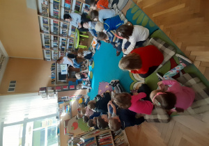 Dzieci siedzą w kole na poduszkach i słuchają książki czytanej przez bibliotekarkę.
