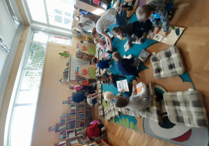 Dzieci oglądają książki dostępne w bibliotece. Niektóre dzieci siedzą na niebieskim dywanie a inne szukają książek na półkach.