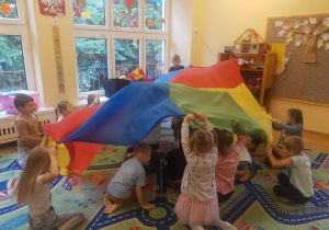 Dzieci zgrupy czerwonej siedzą na dywanie trzymają nad sobą kolorową chustę animacyjną, poruszają nią
