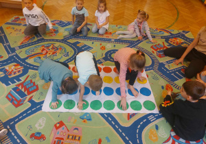 Troje dzieci bawi się na macie twister pozstałe dzieci siedzą na dywanie