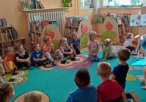 Dzieci z grupy zielonej podczas zajęć bibliotecznych, dzieci siedzą na dywanie i słuchają pani bibliotekarki, która demonstruje czytaną książkę
