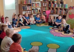 Dzieci z grupy zielonej podczas zajęć bibliotecznych, dzieci siedzą na dywanie i słuchają pani bibliotekarki