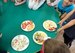Na zdjęciu dzieci z grupy fioletowej siedzą przy stole, na którym znajdują się kawałki owoców i warzyw, jedno z dzieci ma założoną przepaskę na oczy próbuje owoce