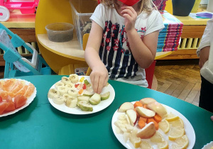 Na zdjęciu dzieci z grupy fioletowej siedzą przy stole, na którym znajdują się kawałki owoców i warzyw, jedno z dzieci ma założoną przepaskę na oczy próbuje owoce