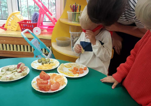 Na zdjęciu dzieci z grupy fioletowej siedzą przy stole, na którym znajdują się kawałki owoców i warzyw, jedno z dzieci ma zakładaną opaskę przez Panią nauczycielkę