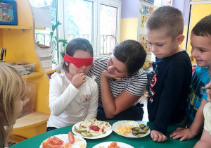 Na zdjęciu dzieci z grupy fioletowej siedzą przy stole, na którym znajdują się kawałki owoców i warzyw, jedno z dzieci ma założoną przepaskę na oczy próbuje owoce obok siedzi Pani nauczycielka