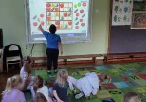 Na zdjęciu dzieci z grupy zielonej podczas zajęć z kodowania jeden z chłopców stoi obok tablicy multimedialnej i wykonuje zadanie z kodowania warzyw