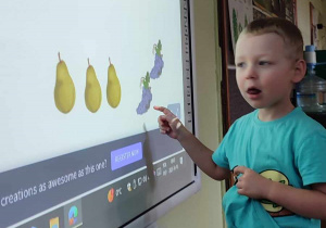 Na zdjęciu chłopiec stoi obok tablicy multimedialnej i wykonuje zadanie układa rytm z owoców