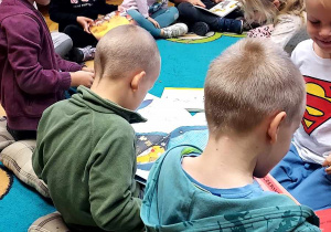 Na zdjęciu dzieci z grupy fioletowej siedzą na dywanie w Bibliotece Miejskiej i oglądają książki