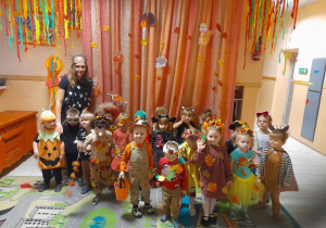 Na zdjęciu dzieci z grupy żółtej wraz z nauczycielem dzieci stoją na dywanie na tle dekoracji jesiennej dzieci są przebrane stroje o tematyce jesiennej
