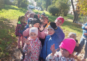 Dzieci ustawione parami są na spacerze w pobliżu przedszkola. Uśmiechają się i machają rękami.
