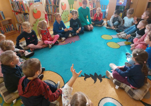 Dzieci siedzą w kole na błękitno-beżowym dywanie. Słuchają opowiadania bibliotekarki o sposobie korzystania z biblioteki.