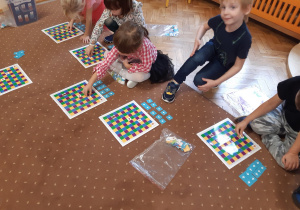 Dzieci siedzą na brązowym dywanie. Układają za pomocą białych strzałek na niebieskim tle drogę, którą odbywa wiewiórka na planszy złożonej z kolorowych kwadratów.