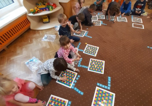 Dzieci siedzą w kole na brązowym dywanie. Przed nimi leżą plansze złożone z kolorowych kwadratów oraz białe strzałki na niebieskim tle służące do kodowania.
