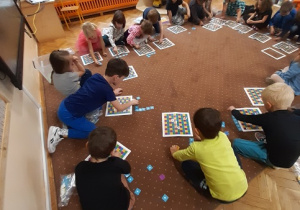 Dzieci siedzą w kole na brązowym dywanie. Pochylają się nad planszami złożonymi z kolorowych kwadratów.