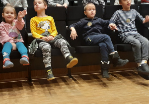 Trzech chłopców i jedna dziewczynka siedzi na fotelach i czekają na rozpoczęcie koncertu w wykonaniu uczniów Szkoły Muzycznej. Dziewczynka macha ręką.