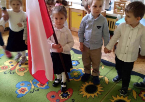 Na zdjęciu dzieci z gruppy żółtej podczas obchodów poprzedzających święto 11 listopada czwórka dzieci stoi na dywaie dziewczynka trzyma w ręku flagę Polski