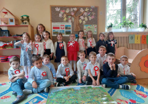 Dzieci w strojach galowych z przyczepionymi kotylionami w biało-czerwonych barwach pozują do zdjęcia. Na dywanie przed dziećmi leży mapa Polski.