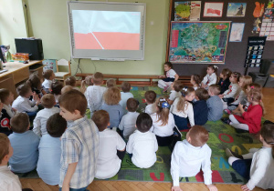 Dzieci siedzą na dywanie. Na tablicy multimedialnej widnieje biało-czerwona flaga Polski.